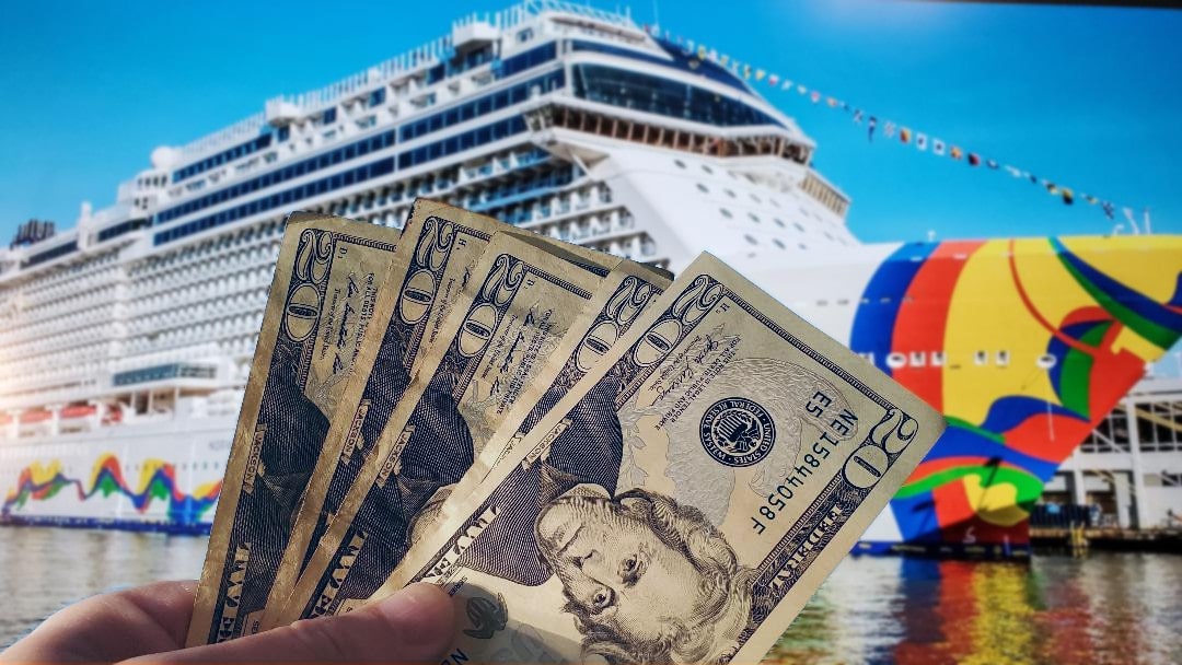 stack of 20 dollar bills in front of a norwegian cruise line cruise ship